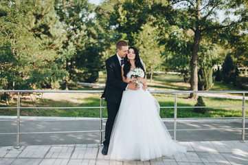 Naklejka premium Beautiful brunette bride in a white dress with a bouquet and stylish groom in a black suit cuddling in the garden with greenery. Portrait of lovers and smiling newlyweds. Wedding photography.