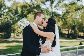 Beautiful brunette bride in a white dress with a bouquet and stylish groom in a black suit cuddling...