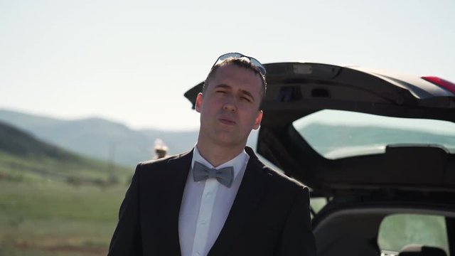 A young man in a classic suit, bow tie smokes VAPE. Stands next to the car in nature.