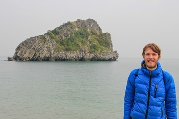 Fototapeta premium A young white attractive male blond with a beard in a blue down jacket stands against the backdrop of a cliff and the sea in Ha Long Bay, Vientam