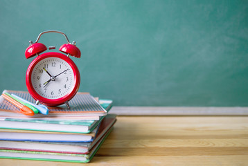 Red alarm clock, books and pens against the background of a green blackboard. School background with space for your text