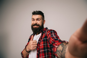 smiling bearded man in checkered shirt showing thumb up while taking selfie, isolated on grey