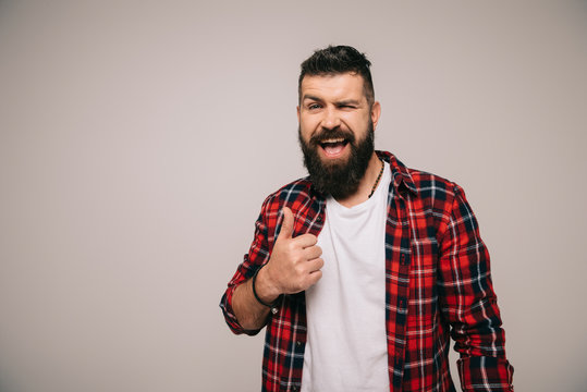 excited bearded man winking and showing thumb up, isolated on grey
