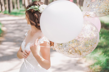 Beautiful young bride holding several air balloons in hands while standing outside on sunny warm...