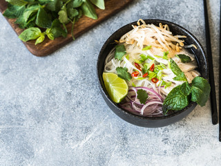 Traditional Vietnamese soup- pho ga in black bowl with chicken and rice noodles, mint and cilantro, green and red onion, chili, bean sprouts and lime on grey background. Asian food.