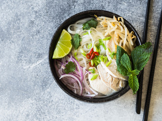 Traditional Vietnamese soup- pho ga in black bowl with chicken and rice noodles, mint and cilantro, green and red onion, chili, bean sprouts and lime on grey background. Asian food. - obrazy, fototapety, plakaty