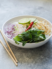 Traditional Vietnamese soup- pho ga in white bowl with chicken and rice noodles, mint and cilantro, green and red onion, chili, bean sprouts and lime on grey background. Asian food. Copy space