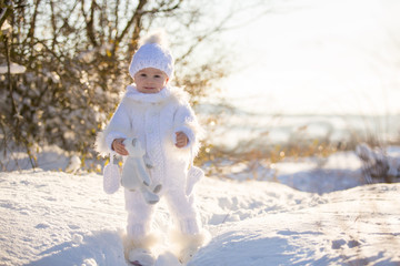 Baby playing with teddy in the snow, winter time. Little toddler boy in handmade white snowsuit, holding teddy bear on sunset, playing outdoors in winter park