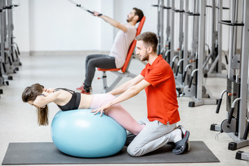 Young woman stretching on a fitness ball with man trainer during the spine treatment at the rehabilitation gym
