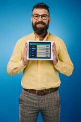 smiling bearded man showing digital tablet with sport app, isolated on blue