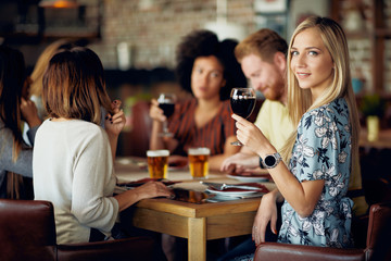 Woman looking at camera and holding glass of wine while sitting in restaurant. In background friends drinking and chatting.