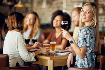 Woman looking at camera and holding glass of wine while sitting in restaurant. In background friends drinking and chatting.