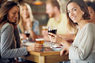 Woman looking at camera and holding glass of wine while sitting in restaurant. In background friends drinking and chatting.