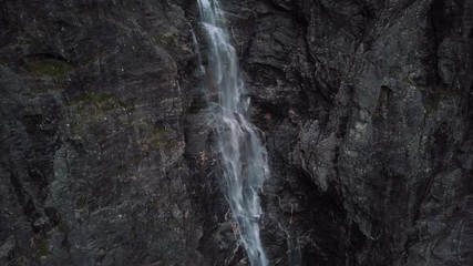 Drone panning around a dramatic waterfall cascading down a black rock cliff face leading to a small river and winding road in Norway