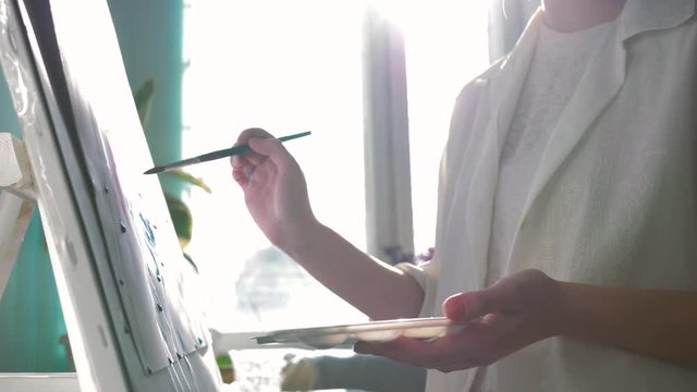 fine art, happy artist female with inspiration relish paints painting with bright colors on white canvas on easel at workroom against sunlit window, shooting in motion