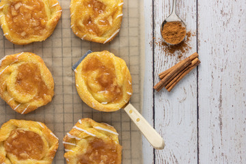 apple danish on cooling rack and parchment paper with spatula cinnamon in a spoon and cinnamon sticks from above on a rustic white wood background