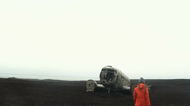 Young traveling woman walking alone on the black beach near the breaker DC-3 BMC plane in Iceland in windy day.