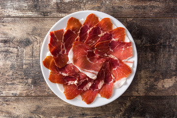 Spanish serrano ham or prosciutto in plate on wooden table on wooden table. Top view