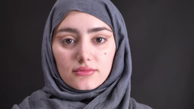 Portrait of young muslim woman in hijab watching calmly in camera on black background.