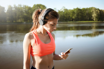 Outdoor pcture of charming self determined young Caucasian woman runner wearing sports top and headphones, holding cell phone, choosing playlist for cardio workout in park with lake behind her