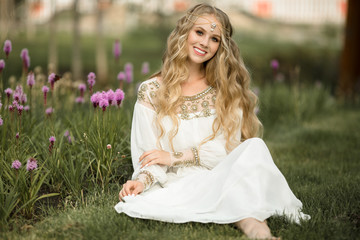 Happy smiling blonde girl is sitting on grass in spring park with purple flowers