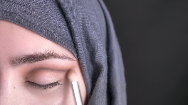 Close-up portrait of female hands doing eye make-up stumping with brush for beautiful muslim woman in hijab on black background.