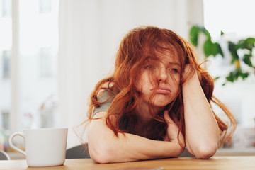 Red-haired girl sitting at table with boring face - obrazy, fototapety, plakaty