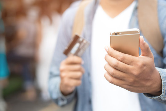 Close Up Young Man Hands Holding Paying With Credit Card And Using Cell, Smart Phone For Online Shopping Instead Of Using Money Standing In Front Of A Clothing Shop Store. 