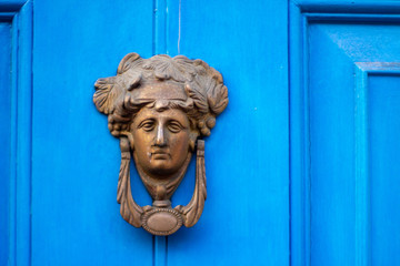 Bronze door knocker in the shape of a woman's head with elaborate hair on a blue house door