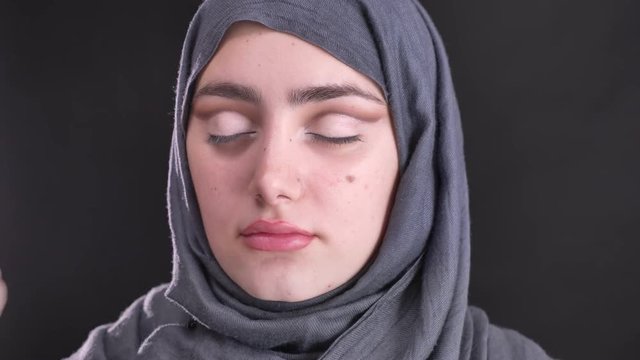 Portrait of female hands doing eye-makeup with thin brush for beautiful muslim woman in hijab on black background.