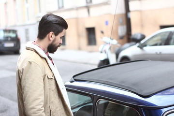 Fototapeta na wymiar The photo of a face of a handsom bearded man gay in brown jacket on the street outside near a blue car