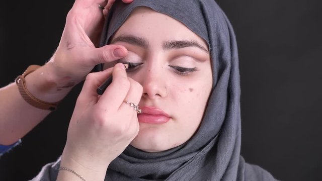 Portrait of female hands doing eye-makeup with black pencil for beautiful muslim woman in hijab on black background.