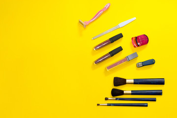 Woman's accessories on yellow background: disposable pink razor, nail file, lipstick, eyelash curler, clippers, brushes of different sizes. Frame of tools for makeup with copy space.
