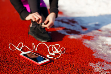 girl athlete in black sneakers, crouching on the red track for running. near is a phone with wired headphones. Cold snowy weather