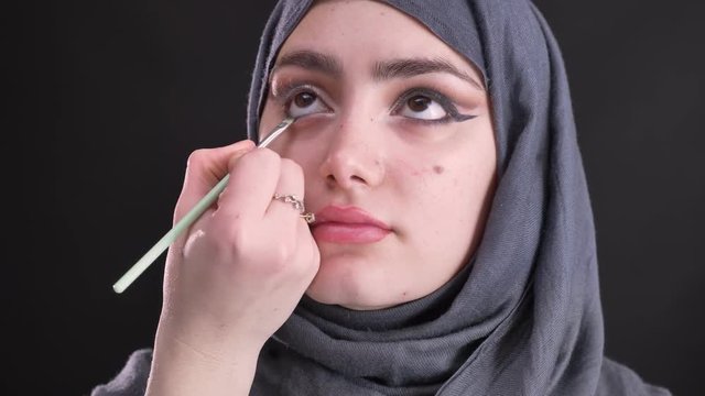 Portrait of female hands drawing black arrow using thin flat brush for attractive muslim woman in hijab on black background.