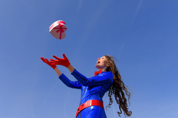 A young woman dressed in shiny blue throws a decorative red heart  shaped gift in the air. She celebrates Valentine's Day and her love to her partner.