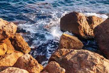 Blue foaming waves beat against large warm stones lit by sun on coast
