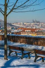 Prague, Czech Republic / Europe - February 5 2019: Scenic view of Prague city and Prague castle from Vysehrad overlooking buildings, ground covered with snow, benches, blue sky, vertical image