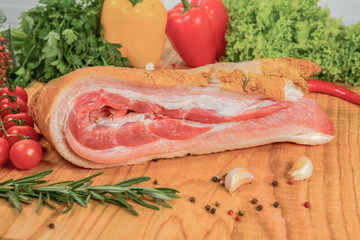 Fresh bacon - lard on a wooden board, with tomatoes on the side, salad and red pepper from behind, rosemary and cloves of garlic in the foreground