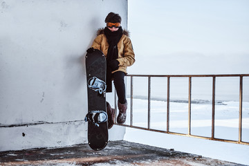 A teenage snowboarder dressed in warm clothes and protective goggles sitting on railing near the snowy beach