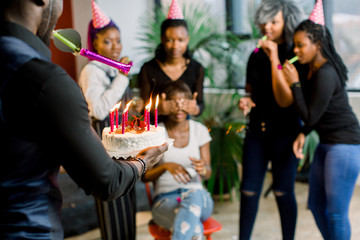 Attractive young african woman with dark hair sitting on the chair with her friends around her, celebrating and blowing out her birthday candles. An African guy carries birthday cake with candles