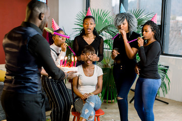 Attractive young african woman with dark hair sitting on the chair with her friends around her, celebrating and blowing out her birthday candles. An African guy carries birthday cake with candles