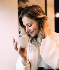 blonde girl in a cozy warm cafe, in a white knitted sweater drinking coffee.