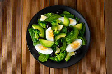 Avocado salad with broccoli, spinach, olives and boiled eggs in black plate, over wooden table. Healthy food. Avocado salad recipe.
