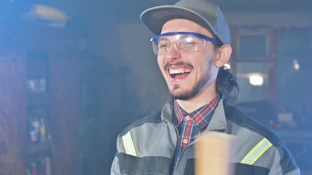 Concentrated young bearded joiner in goggles and a cap checks the workpiece for suitability while holding it in his hands and examining