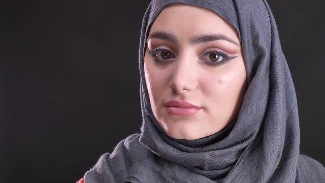 Portrait in profile of beautiful muslim woman in hijab with fashionable make-up turns to camera and watches calmly on black background.