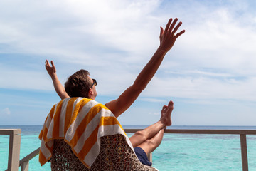 young man in swimsuit relaxing on a terrace and enjoying freedom in a tropical destination. Arms...