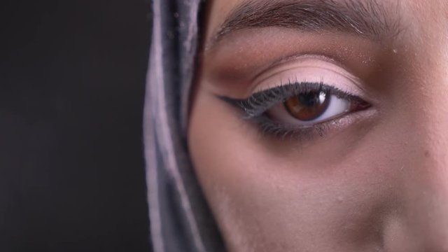 Close-up half-portrait of young muslim woman in hijab with fashionable make-up watching downwards on black background.
