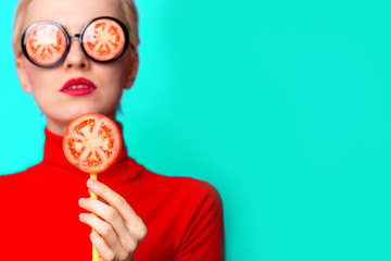 Amazing cute young pretty girl on the turquoise background wearing sunglasses with tomato and holding lollipop . Blond sex model,  trendy glamour look, stylish party look