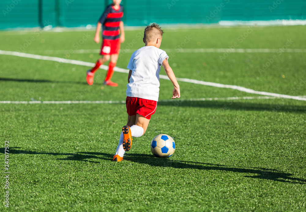 Wall mural Boys in red white sportswear running on soccer field. Young footballers dribble and kick football ball in game. Training, active lifestyle, sport, children activity concept 